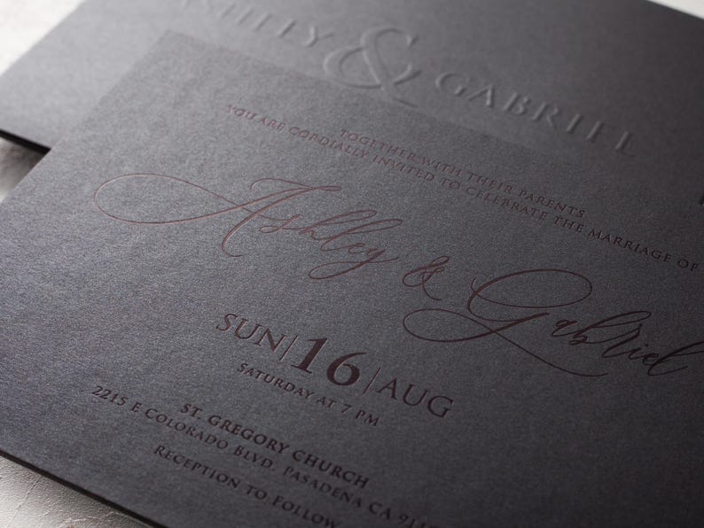 A close-up of a black wedding invitation with "Ashley & Gabriel" embossed in copper script, along with the date "SUN 16 AUG" and event details, on a textured surface.