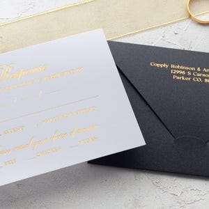 A white RSVP card with gold foil text and a black envelope with a printed return address are shown on a light gray surface, with a gold ribbon and two gold rings.