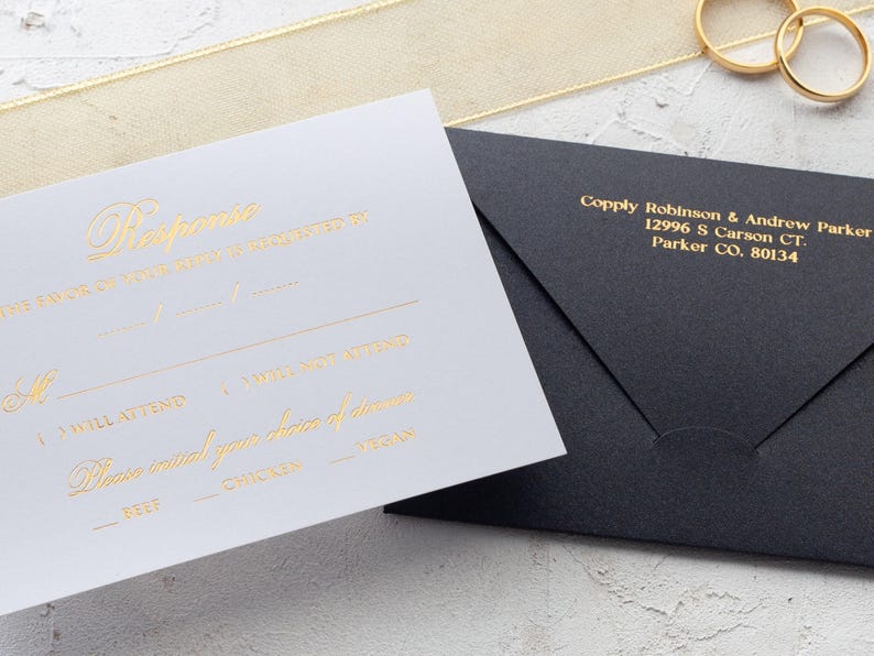 A white RSVP card with gold foil text and a black envelope with a printed return address are shown on a light gray surface, with a gold ribbon and two gold rings.