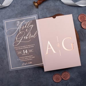 Personalized clear acrylic wedding invitation beside a blush pink sleeve envelope with rose gold foil monogram.