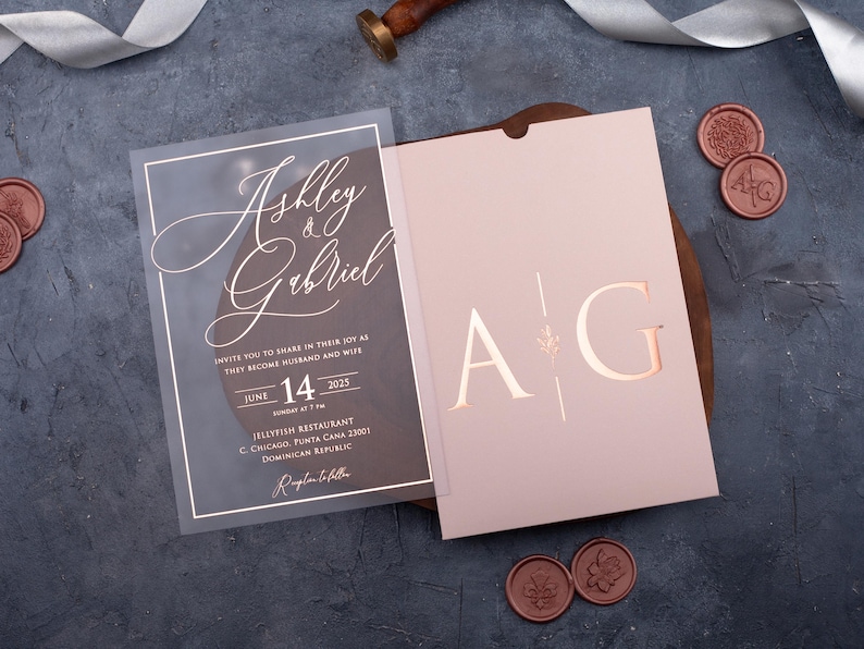 Personalized clear acrylic wedding invitation beside a blush pink sleeve envelope with rose gold foil monogram.