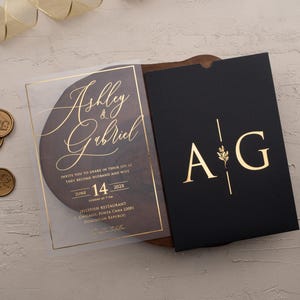 Personalized clear acrylic wedding invitation with rose gold foil beside a black sleeve envelope with monogram.