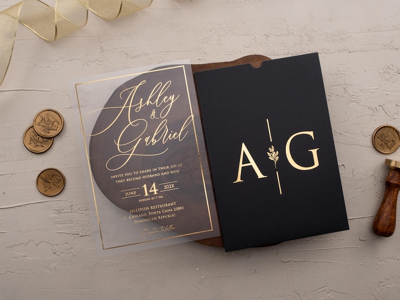 Personalized clear acrylic wedding invitation with rose gold foil beside a black sleeve envelope with monogram.
