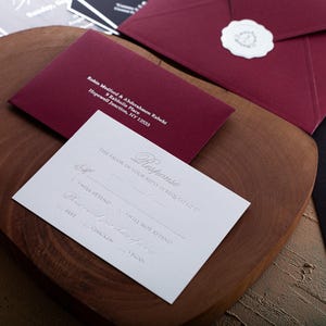A white RSVP card with embossed text and a dark red envelope with a printed address and a wax seal are displayed on a wooden board.