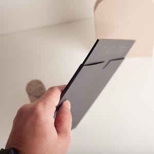 A hand holds a black wedding invitation card with embossed text, partially inserted into a light-colored slot on a shelf.
