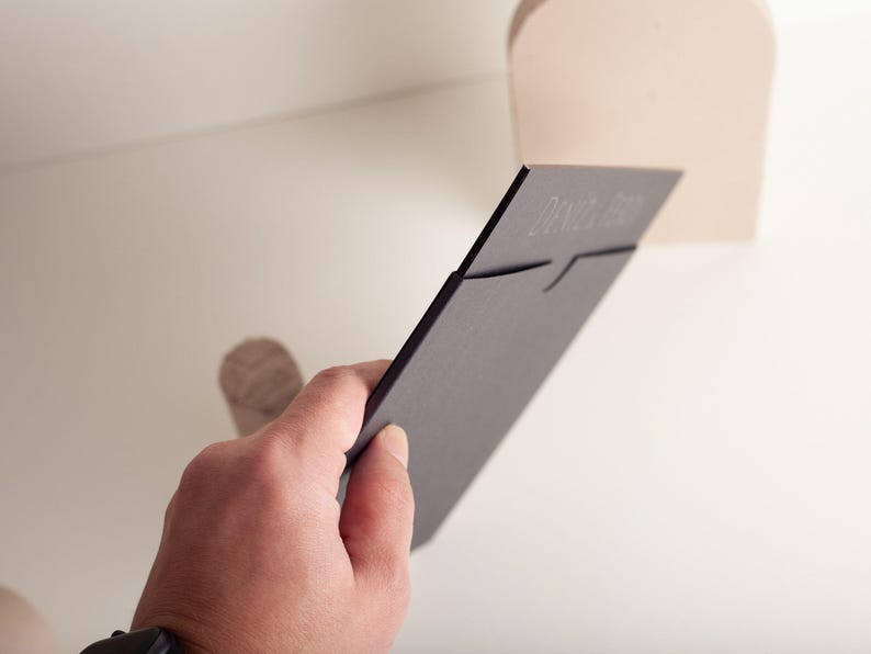 A hand holds a black wedding invitation card with embossed text, partially inserted into a light-colored slot on a shelf.