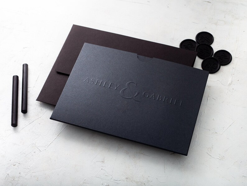 A black wedding invitation with embossed names on a black envelope and a black card are arranged on a white textured surface, with black wax seals and wax sticks nearby.