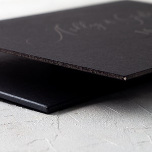 May include: Close-up of two black rectangular objects, possibly a book or a sign, with a textured edge. The top object has faint, handwritten text. The objects are on a white, textured surface.