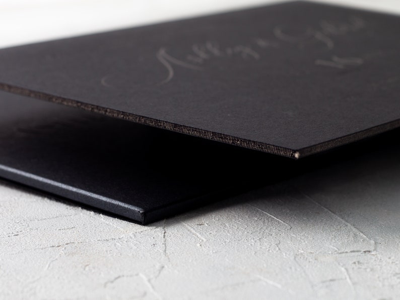 May include: Close-up of two black rectangular objects, possibly a book or a sign, with a textured edge. The top object has faint, handwritten text. The objects are on a white, textured surface.