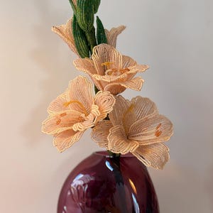 May include: A handcrafted beaded flower stem featuring pale peach petals and green beaded leaves, displayed in a rounded, deep purple glass vase with an embossed floral pattern on its surface. The flower has delicate stamens and textured beads.