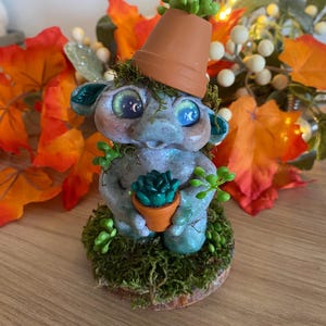 May include: A whimsical blue and green gnome figurine with large eyes, holding a small potted succulent. The gnome is wearing a terracotta pot as a hat and is sitting on a mossy base.