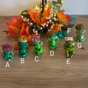 May include: Seven green frog figurines with glitter accents, each wearing a different colored crown. The frogs are sitting on a wooden surface with fall foliage in the background.