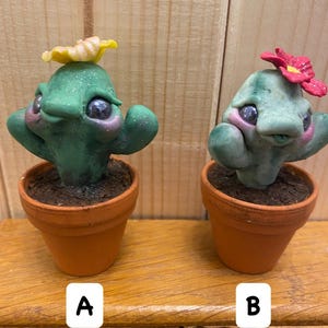 May include: Two small potted cactus plants with painted faces and flowers on top. The cactus on the left has a yellow flower and the cactus on the right has a red flower. Both are in terracotta pots.