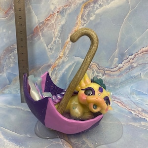 May include: A whimsical sculpture of a yellow dragon nestled inside a purple and lavender umbrella. The umbrella handle is gold glitter. The dragon has purple eye makeup and a blue gem. The sculpture is approximately 6 inches tall.