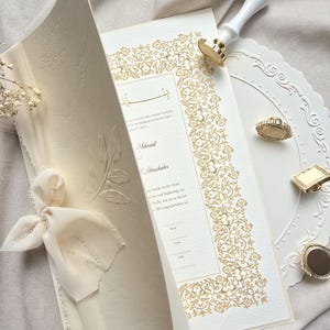 Op de afbeelding: Ivoor trouwkaart met gouden bloemenrand en tekst. Een bijpassende lakstempel en decoratieve ring liggen op een wit bord. De uitnodiging is vastgebonden met een crèmekleurig lint en strik. Het thema is elegant en formeel.