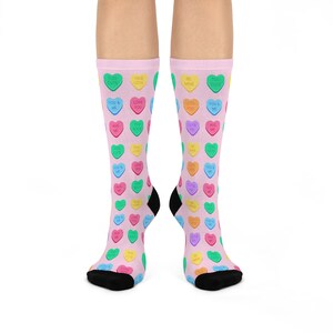 Pode incluir: Um par de meias cor-de-rosa com um padrão de corações de doces coloridos. Os corações têm várias mensagens, incluindo "Love You", "Hug Me", "You & Me" e "Cutie Pie".