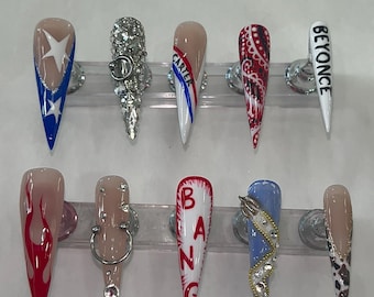 May include: A collection of ten long, almond-shaped artificial nails. Designs include a white star on blue, silver rhinestones, red and white stripes, a paisley pattern, the word "BEYONCE", flames, a silver ring, the word "BANG", a blue nail with gold accents, and a cow print.