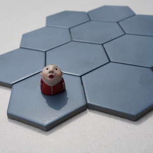 Puede incluir: Una pequeña figurita de cerámica de una persona con cara redonda y camisa roja, sentada en un azulejo hexagonal azul.