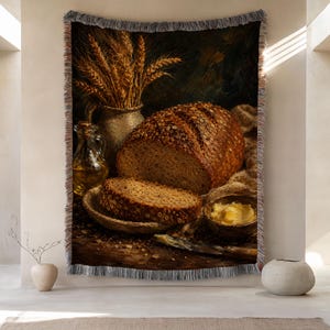 May include: A woven throw blanket featuring a still life print of a loaf of seeded bread, a slice of bread on a plate, a small bowl of butter, a glass bottle of oil, and a pitcher of wheat stalks, finished with decorative fringe on all edges.