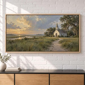 May include: A landscape painting in a wooden frame depicting a white church with a steeple situated in a grassy field near a river at sunset. The sky features soft yellow and blue clouds, while large trees with hanging moss frame the scene.