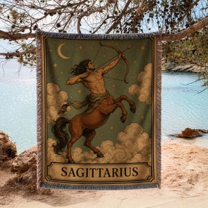 May include: A rectangular woven throw blanket featuring a central illustration of a centaur archer aiming a bow and arrow. The design includes a crescent moon, stars, and clouds against a sage green background, with the text SAGITTARIUS at the bottom.