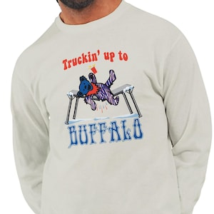 May include: Cream-colored long-sleeve shirt with a graphic featuring a blue bear with a purple and white striped body, lying on a snow-covered table. The text "Truckin' up to" is above the bear, and "BUFFALO" is below.