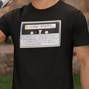 May include: Black t-shirt with a white cassette tape graphic. The tape label reads "NY MSG - 10/12/83" and lists the setlist for a concert. The setlist includes "Cold Rain and Snow", "Mingewand Blues Ramble on Rose", "My Brother Esau", "It Must Have Been the Roses", "cansay converting lies", "Looks Like Rams might as well", "Help Sipbret Franks Man", "Smart Woman Smarter", "Hes lamer Drums Space", "Trick Back Peter", "Not Fade Away", "ticore Revolition".