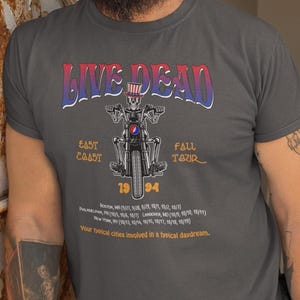 May include: A dark gray t-shirt with a graphic of a skeleton riding a motorcycle. The graphic is in a vintage style with the words "Live Dead" in a colorful font. The text "East Coast Fall Tour 1994" is also included on the shirt. The shirt has a crew neck and short sleeves.