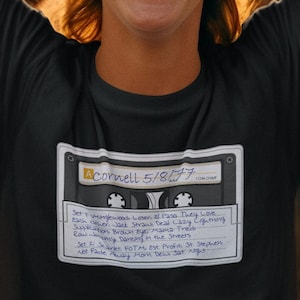 May include: A black t-shirt with a white cassette tape graphic. The tape label reads "A Cornell 5/8/77" and lists the songs "Minglewood Loser", "El Paso", "They Love Each Other", "Jack Straw", "Deal Lazy Lightning", "Supplication", "Brown Eye Mama Fried", "Row Jimmy", "Dancing in the Streets", "Scarlet", "FOTM est. Profit", "St. Stephen", and "Not Fade Away Morn Dew Sat. Night."