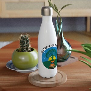 May include: White insulated water bottle with a silver cap. The bottle features a circular graphic with the text "DAYBREAK ON THE" and an illustration of a tree and a character. The bottle is on a wooden coaster.