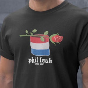 May include: Black t-shirt with a red, white, and blue design of a can with a rose on top. The text "phil lesh 1940-2024" is printed below the design.