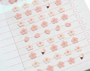 May include: A white notepad featuring a grid of light pink horizontal lines and a vertical column of empty checkboxes on the right side. The page is decorated with numerous small, die-cut pink cherry blossom stickers with gold centers.