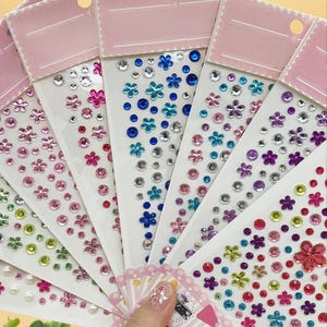 May include: A collection of decorative sticker sheets featuring various floral and gem designs. The stickers come in a variety of colors, including pink, blue, purple, and clear, with a mix of flower and round shapes. Each sheet has a pink header.
