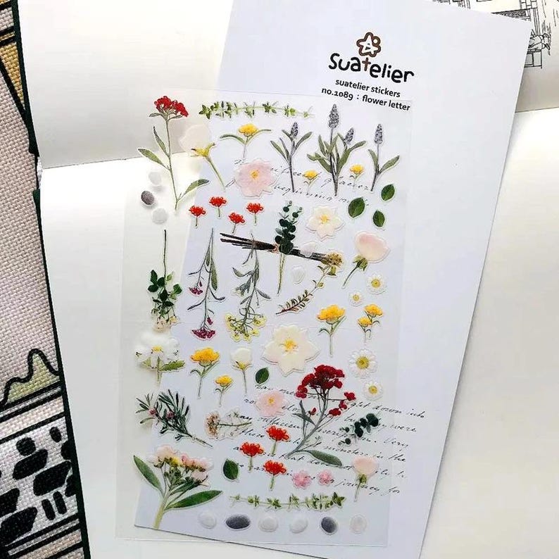May include: A clear sheet of Suatelier stickers no.1089 flower letter featuring various botanical illustrations including red, yellow, and white flowers, green stems, leaves, and small pebbles, arranged on a white backing card with cursive text.