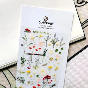 May include: A sheet of Suatelier stickers no.1089 flower letter featuring various botanical illustrations including red, yellow, white, and pink flowers, green leaves, and stems on a clear backing. The header displays the brand name and product details.