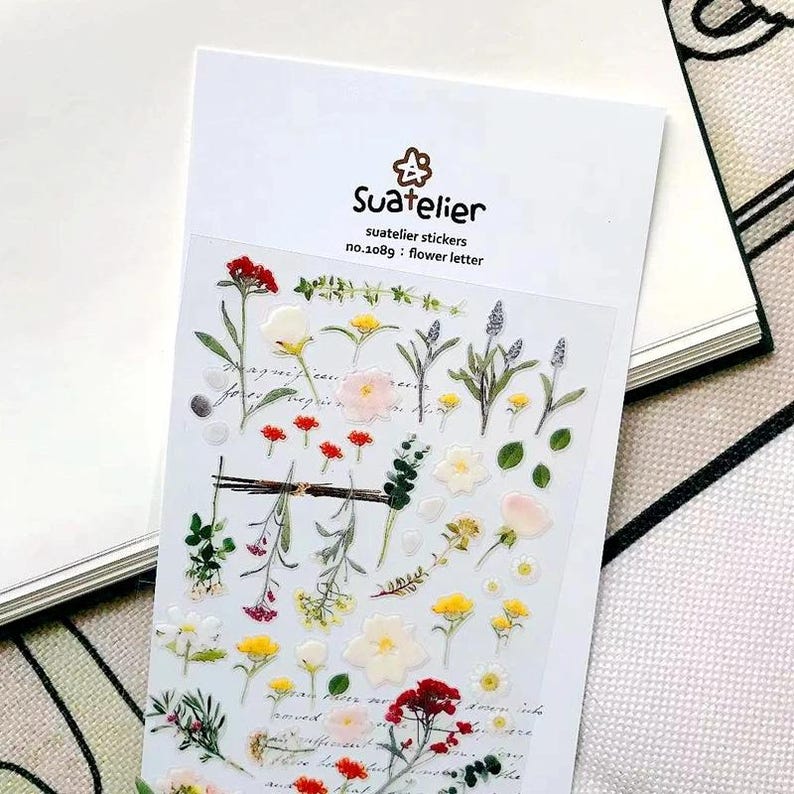 May include: A sheet of Suatelier stickers no.1089 flower letter featuring various botanical illustrations including red, yellow, white, and pink flowers, green leaves, and stems on a clear backing. The header displays the brand name and product details.