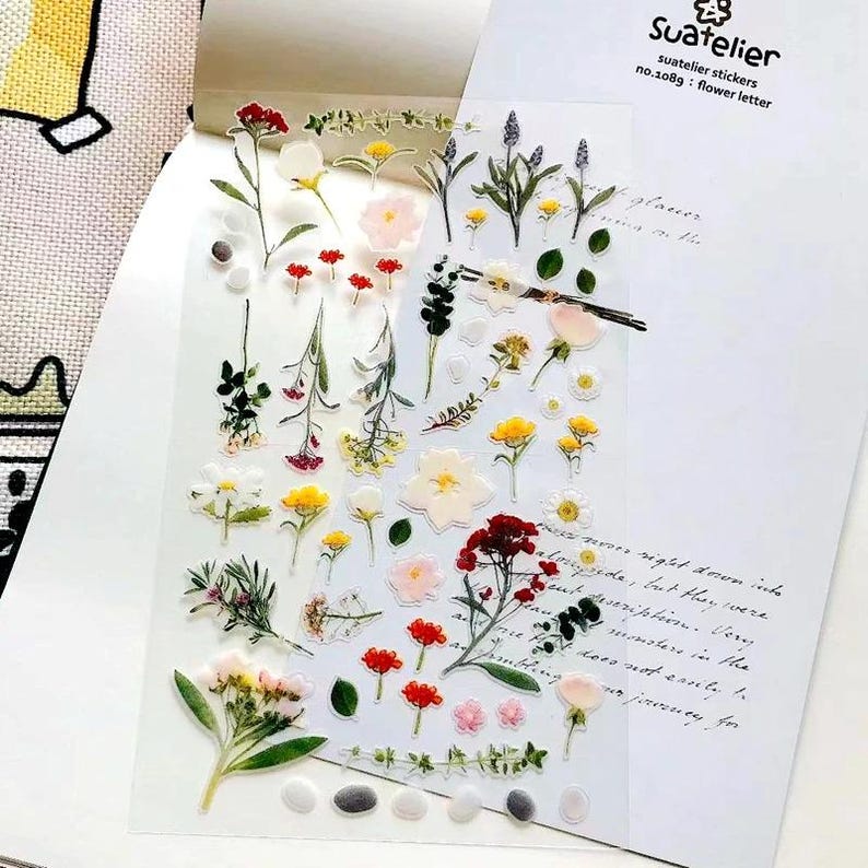 May include: A clear sticker sheet labeled Suatelier no.108g flower letter featuring various botanical illustrations including red, yellow, and white flowers, green leaves, sprigs, and small gray pebbles arranged on a white backing paper.