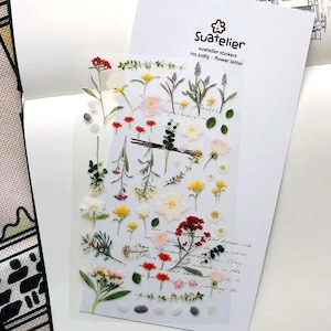 May include: A clear sheet of Suatelier stickers no.1089 flower letter featuring various botanical illustrations including red, yellow, and white flowers, green stems, leaves, and small pebbles, arranged on a white backing card with cursive text.