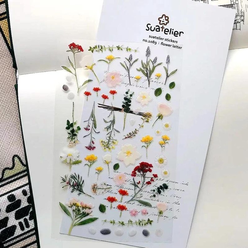 May include: A clear sheet of Suatelier stickers no.1089 flower letter featuring various botanical illustrations including red, yellow, and white flowers, green stems, leaves, and small pebbles, arranged on a white backing card with cursive text.