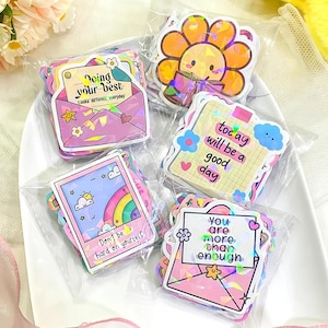 Puede incluir: Una colección de pegatinas coloridas en envases de plástico transparente. Las pegatinas presentan varios diseños, incluyendo un sobre con el texto "Doing your best", un arcoíris, un girasol y el texto "You are more than enough".