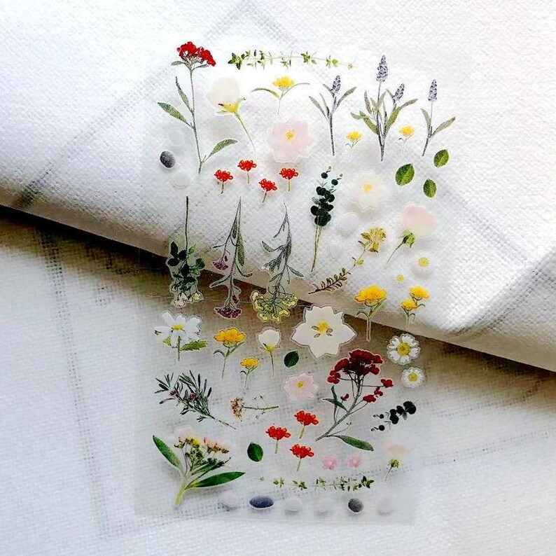 May include: A clear sticker sheet featuring a variety of botanical illustrations including red, yellow, white, and green wildflowers, herbs, and small leaves. The designs are arranged in a scattered pattern across the transparent rectangular sheet.