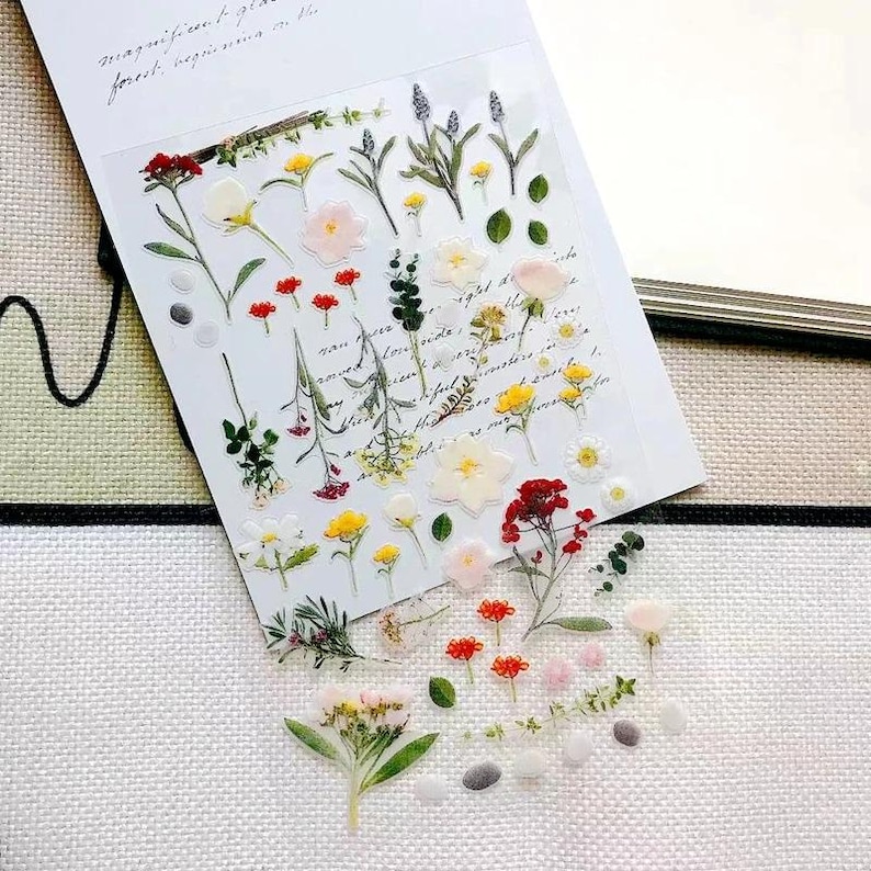 May include: A clear sticker sheet featuring a variety of botanical illustrations including red, yellow, white, and pink flowers, green leaves, and sprigs. The sheet is overlaid on a white background with cursive script text visible beneath the stickers.