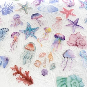 Puede incluir: Una colección de pegatinas estilo acuarela con diversas criaturas marinas. Las pegatinas incluyen medusas, estrellas de mar, conchas y coral en tonos de azul, rosa, morado y amarillo. Las pegatinas están dispuestas sobre una superficie blanca texturizada.