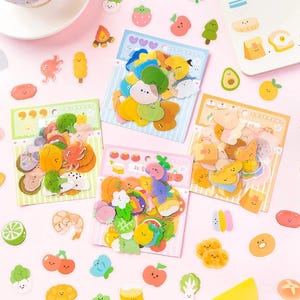 May include: A collection of colorful food-themed stickers, including fruits, vegetables, and other snacks. The stickers are arranged on pink and white surfaces, with some packaged on backing cards. The designs feature cute, cartoon-style illustrations.