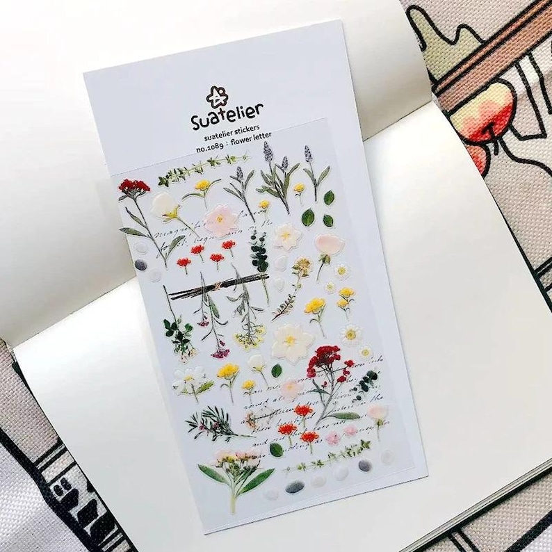 May include: A clear sheet of Suatelier stickers, no. 1089 flower letter, featuring various botanical illustrations including red, yellow, and white flowers, green leaves, and stems, with cursive script text printed in the background of the sticker sheet.