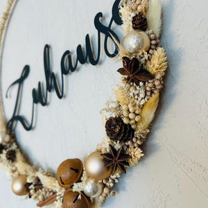 Op de afbeelding: Een cirkelvormige krans met het woord "Zuhause" in zwart script. De krans is versierd met gedroogde bloemen, dennenappels, steranijs en gouden en zilveren ornamenten. De krans is gemaakt van een geweven touwmateriaal.