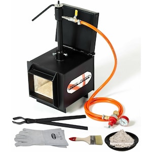 Puede incluir: Un horno de metal negro con una manguera naranja y una etiqueta roja y blanca que dice "Devil-Forge". El horno está sobre un fondo blanco e incluye un par de tenazas, un par de guantes de cuero, un pincel y un pequeño plato de mortero refractario.