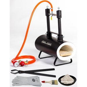 May include: A black metal forge with an orange hose, a regulator, and a pair of tongs. The forge has a white ceramic lining and a label that reads "DEVIL FORGE". The set also includes a pair of gloves, a paintbrush, and a bag of refractory mortar.