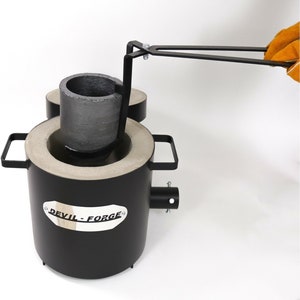 Puede incluir: Un horno de crisol de metal negro con un crisol extra&iacute;ble y un asa. El horno tiene una etiqueta que dice "DEVIL-FORGE".