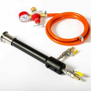 Puede incluir: Un soplete de metal negro con una boquilla de acero inoxidable, dos válvulas de latón con manijas amarillas y una manguera naranja con una conexión de latón. El soplete está conectado a un manómetro con una cara roja y un regulador de latón.